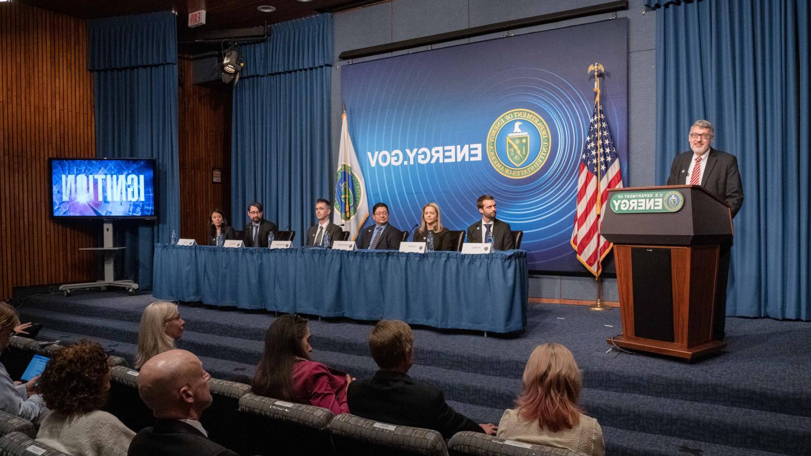 Alex Zylstra '09 and five colleagues are seated at a table; a presenter is standing at a podium during a press conference panel.