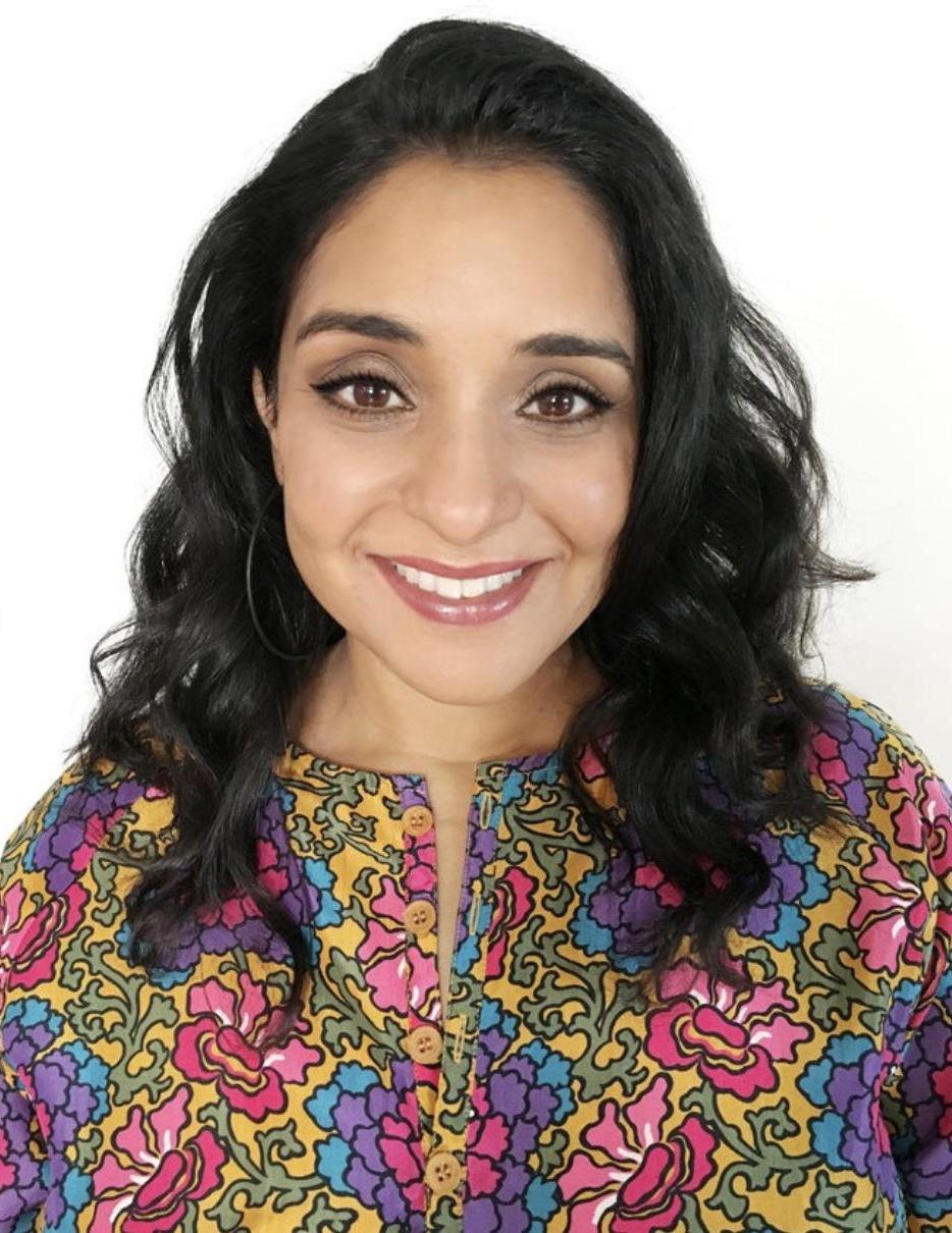 Photo of creative writing workshop leader, local author Neela Banerjee. An Indian-American woman with long, wavy black hair and a bright yellow, purple, blue, pink, and green floral top smiles at the camera.